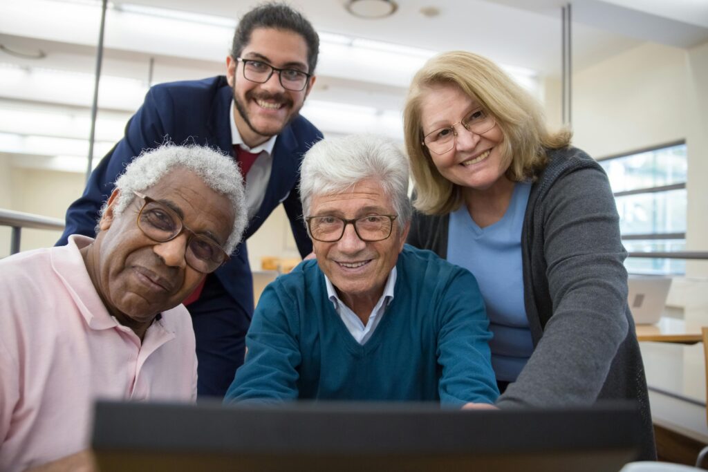 Group of smiling seniors and a young man gathered around a laptop indoors.