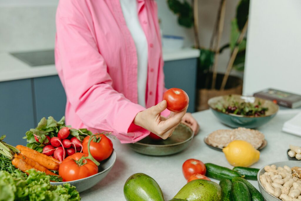 Person in pink overshirt holding a fresh tomato with various organic vegetables on a countertop.