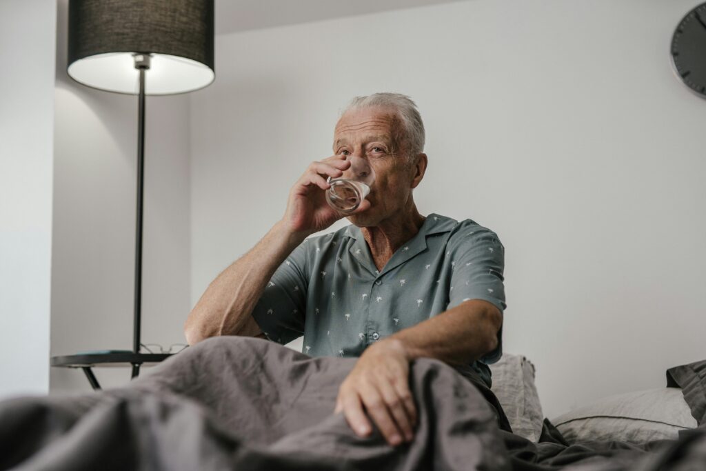 Senior man with gray hair drinks water in bedroom setting, conveying calm and routine.