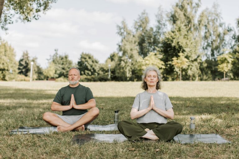 Senior couple meditating on yoga mats in a serene park setting, enjoying peaceful yoga practice.