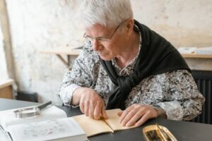 Elderly man with eyeglasses studying at a desk with books and magnifying glass.