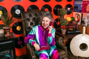 A joyful senior woman in vibrant clothes, posing with a vintage phone, surrounded by vinyl records in a retro-themed room.