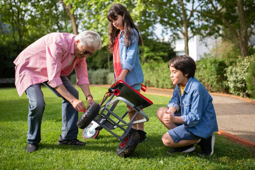 An elderly man assists two children in fixing a garden cart outdoors, showcasing family bonding and teamwork.