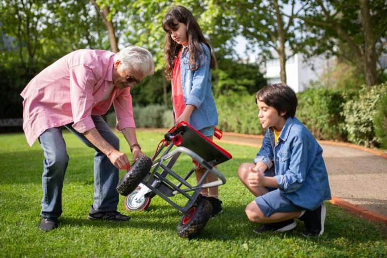 An elderly man assists two children in fixing a garden cart outdoors, showcasing family bonding and teamwork.