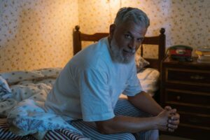 Elderly man with clasped hands sits peacefully on bed in a cozy, softly lit bedroom.