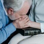 An elderly man expressing sorrow while holding a woman's hand and a photo album, indoors.
