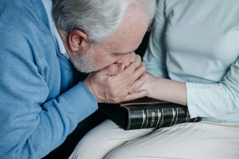 An elderly man expressing sorrow while holding a woman's hand and a photo album, indoors.