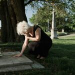 A senior woman in a black dress kneeling at a grave in a tranquil cemetery.