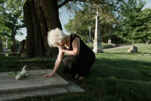 A senior woman in a black dress kneeling at a grave in a tranquil cemetery.