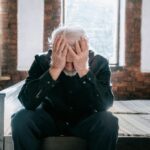 An elderly man sitting on a bed indoors, expressing grief and loss with his head in his hands.