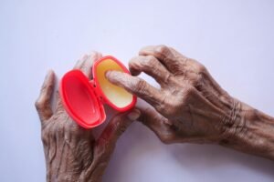 Close-up of senior hands using hand cream for skincare in a red container on a white background.
