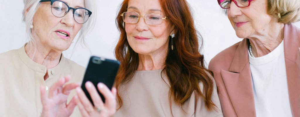 Three senior women enjoying a friendly moment together, sharing a smartphone and smiling.