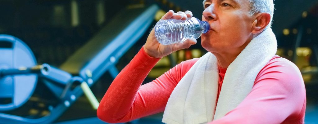 Senior man in gym resting and drinking water post workout for hydration.