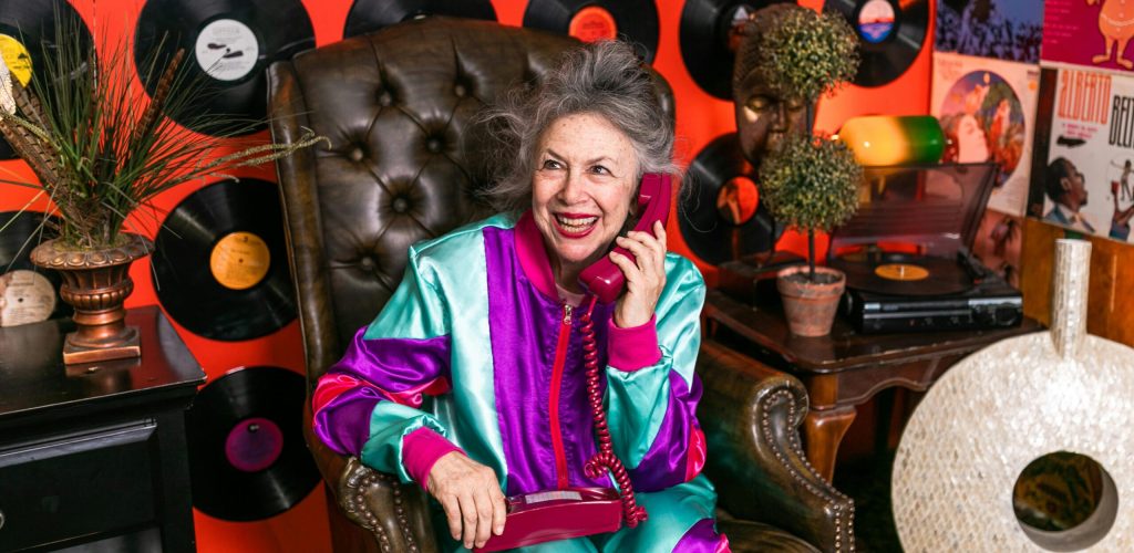 A joyful senior woman in vibrant clothes, posing with a vintage phone, surrounded by vinyl records in a retro-themed room.