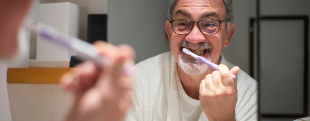 pexels-photo-7477691-7477691 Elderly man in eyeglasses brushes teeth in front of bathroom mirror, smiling warmly.