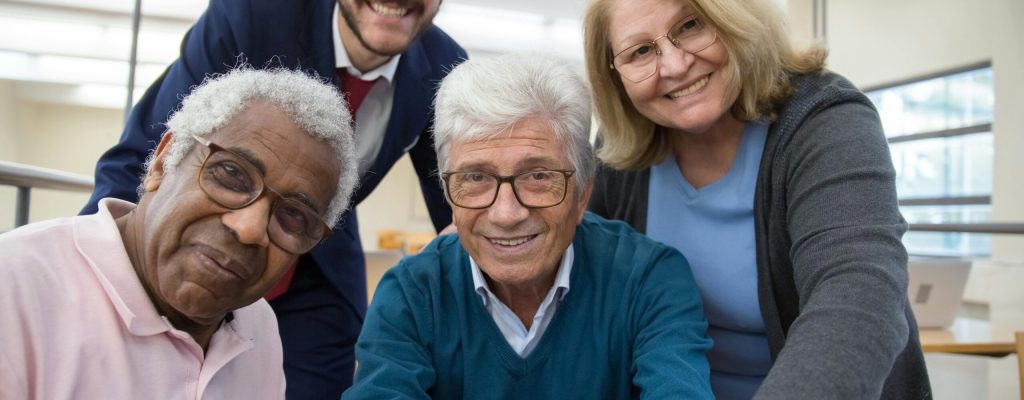Group of smiling seniors and a young man gathered around a laptop indoors.