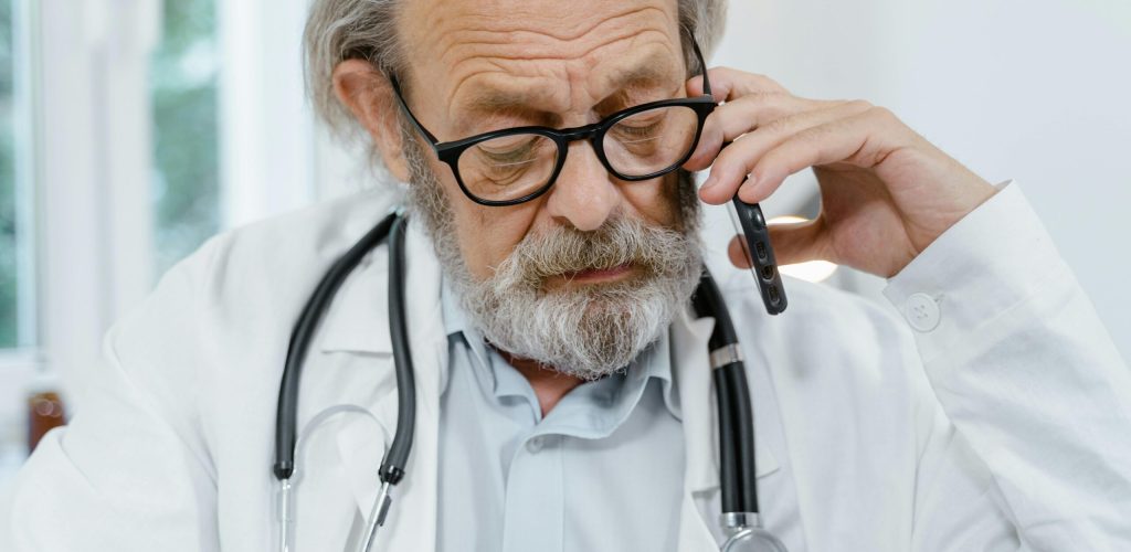 Elderly male doctor in white coat and glasses talking on phone, suggesting telemedicine.