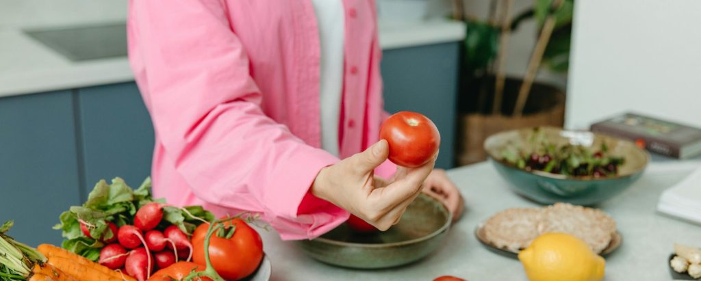 Person in pink overshirt holding a fresh tomato with various organic vegetables on a countertop.